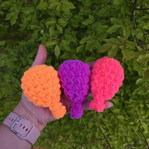 May include: Three handmade crochet balloon-shaped toys in vibrant colors: orange, purple, and pink. The toys are held in a hand against a backdrop of green foliage. The watch on the wrist has a pink strap.