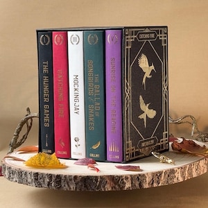 May include: A boxed set of five books from "The Hunger Games" series, displayed on a wooden slice. The books have different coloured spines: black, red, white, green, and purple. The box is dark brown with gold accents and the title "Catching Fire".