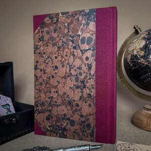 Puede incluir: Un libro de tapa dura con una cubierta veteada marr&oacute;n y negra y un lomo burdeos. El libro est&aacute; de pie sobre una mesa junto a un globo terr&aacute;queo y una peque&ntilde;a caja negra.