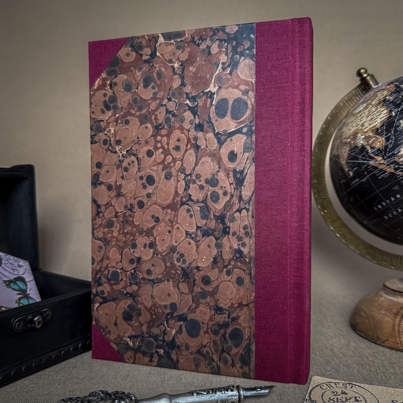 Puede incluir: Un libro de tapa dura con una cubierta veteada marr&oacute;n y negra y un lomo burdeos. El libro est&aacute; de pie sobre una mesa junto a un globo terr&aacute;queo y una peque&ntilde;a caja negra.
