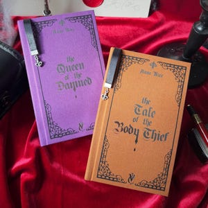 May include: Two hardcover books with black bookmarks and silver charms. The book on the left is purple and titled "The Queen of the Damned" by Anne Rice. The book on the right is brown and titled "The Tale of the Body Thief" by Anne Rice.