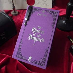 May include: A purple hardcover book titled "The Queen of the Damned" with silver lettering and decorative border. A black bookmark with a silver charm is inserted. The book is on a red velvet surface.