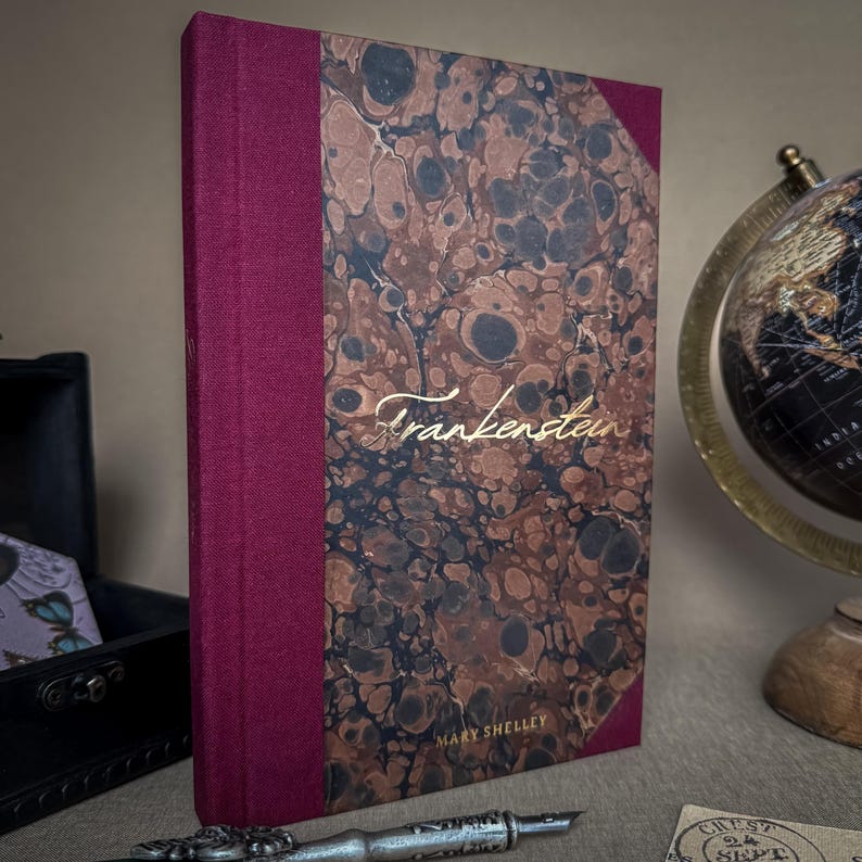 Puede incluir: Un libro de tapa dura titulado "Frankenstein" de Mary Shelley. El libro tiene una cubierta veteada en tonos marrones y negros, con un lomo burdeos. El t&iacute;tulo, en escritura dorada, es visible. El libro est&aacute; sobre una superficie neutra.