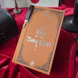 May include: A brown hardcover book with a black bookmark. The book title is "The Tale of the Body Thief" by Anne Rice. The book cover has a decorative border and a silver drop of blood.