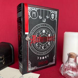 May include: A black book with silver and red accents, featuring the word "Alchemisen" in red script. The cover has a circular design with crescent moons and symbols. A red ribbon bookmark with a charm hangs from the top. The book is on a red surface.