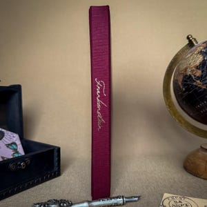 Puede incluir: Lomo de libro de color burdeos con el t&iacute;tulo "Frankenstein" en escritura elegante. El libro est&aacute; en posici&oacute;n vertical, mostrando su t&iacute;tulo. El fondo incluye un globo terr&aacute;queo, un peque&ntilde;o cofre y una pluma decorativa.