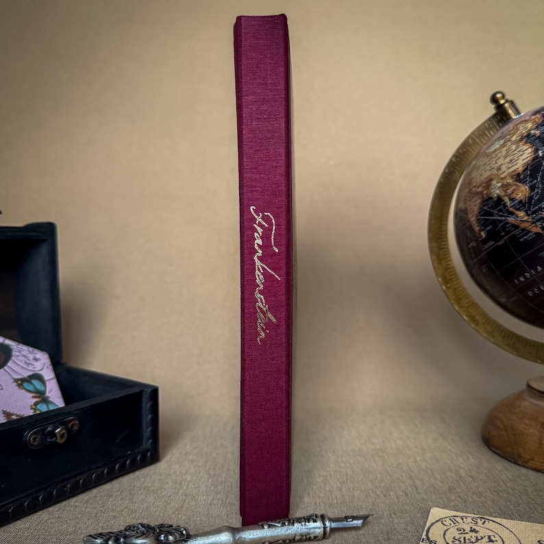 Puede incluir: Lomo de libro de color burdeos con el t&iacute;tulo "Frankenstein" en escritura elegante. El libro est&aacute; en posici&oacute;n vertical, mostrando su t&iacute;tulo. El fondo incluye un globo terr&aacute;queo, un peque&ntilde;o cofre y una pluma decorativa.