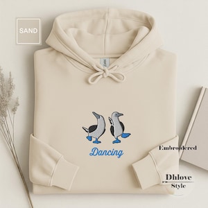 May include: Sand-colored hoodie with an embroidered design of two blue-footed boobies. The word "Dancing" is embroidered in blue below the birds. The hoodie has a drawstring hood and ribbed cuffs.
