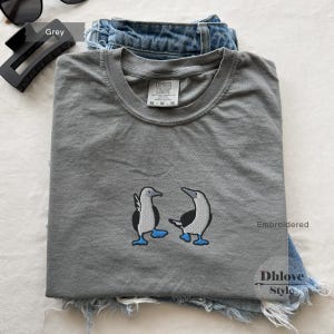 May include: A grey t-shirt with a crew neck featuring an embroidered design of two blue-footed boobies. The shirt is folded on top of a pair of blue denim shorts. The tag reads "Comfort Colors".