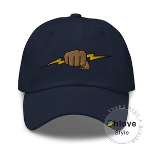 May include: Navy blue baseball cap featuring an embroidered design of a brown fist clenching a yellow lightning bolt. The cap has a curved brim and a structured crown. The embroidered design is centered on the front of the cap.