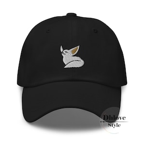 May include: Black baseball cap featuring a white and tan embroidered fox design. The cap has a curved peak and an adjustable strap. The fox design is a stylized illustration.