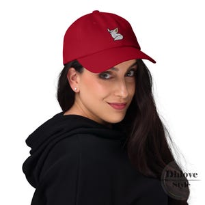 May include: A red baseball cap with a curved peak. The cap has an embroidered design of a white fox with yellow accents on the front. The cap is made of a soft material and has an adjustable strap.