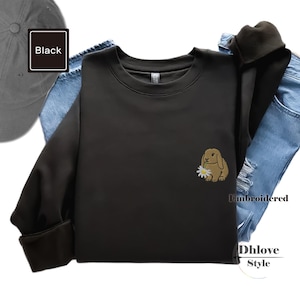 Könnte beinhalten: Schwarzes Sweatshirt mit Rundhalsausschnitt und einem gestickten braunen Hasen, der eine Gänseblümchen hält. Das Sweatshirt wird mit einer blauen Jeans und einer grauen Baseballkappe kombiniert. Das Wort "Black" befindet sich in einer schwarzen Box.