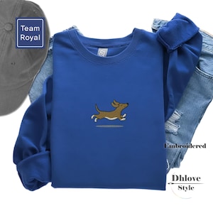 Pode incluir: Um moletom azul royal com um design bordado de cão dachshund marrom. O moletom é combinado com um boné de beisebol cinza e jeans azuis. A palavra "Embroidered" é visível no lado direito da imagem.