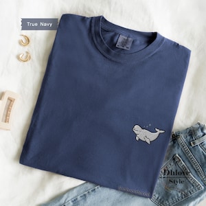 May include: A true navy blue short-sleeve t-shirt featuring a small white embroidered beluga whale graphic on the lower right side. The shirt has a crew neckline and a visible interior tag. The text True Navy and Embroidered are present.