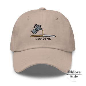 Könnte beinhalten: Beige Baseball Cap mit gebogenem Schirm. Die Kappe hat ein gesticktes Design einer Kaffeemaschine, die Kaffee auf einen Ladebalken gießt, mit dem Wort "LOADING" darunter. Die Kappe hat eine strukturierte Passform.