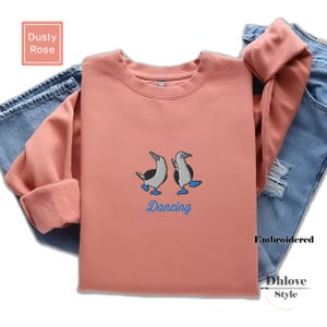 May include: Dusty rose-colored sweatshirt with embroidered blue-footed boobies and the word "Dancing" in blue. The sweatshirt has long sleeves and is paired with blue jeans.