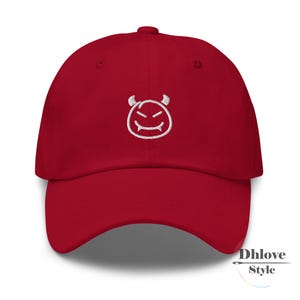 May include: A red baseball cap with a white embroidered devil face on the front. The cap has a curved brim and a fabric strap closure. The text "Dhlove Style" is printed on the lower right side of the cap.