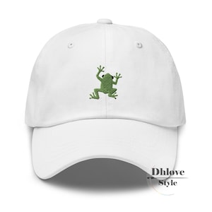 Könnte beinhalten: Weiße Baseballkappe mit einem grünen, gestickten Frosch-Design. Die Kappe hat einen gebogenen Schirm und einen verstellbaren Riemen. Der Frosch ist in einem einfachen, cartoonartigen Stil dargestellt.