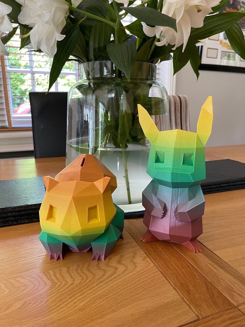 May include: Two colorful, 3D printed figurines of Pokemon characters. The figurine on the left is yellow, orange, and green, and the figurine on the right is green, purple, and pink.