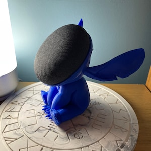 May include: A blue 3D printed Stitch character figurine holding a black smart speaker. The figurine is sitting on a white circular base with a decorative pattern.