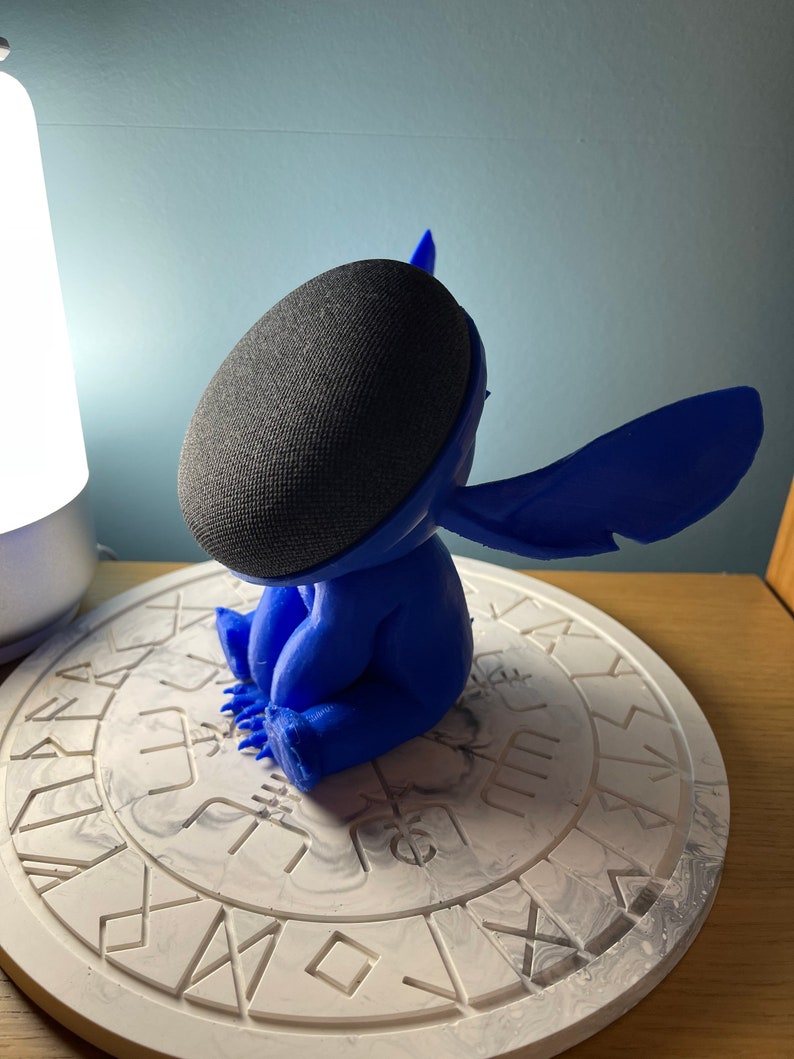 May include: A blue 3D printed Stitch character figurine holding a black smart speaker. The figurine is sitting on a white circular base with a decorative pattern.