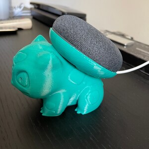 May include: A teal green 3D printed figurine of a Pokemon character, Bulbasaur, holding a gray Google Home Mini smart speaker.