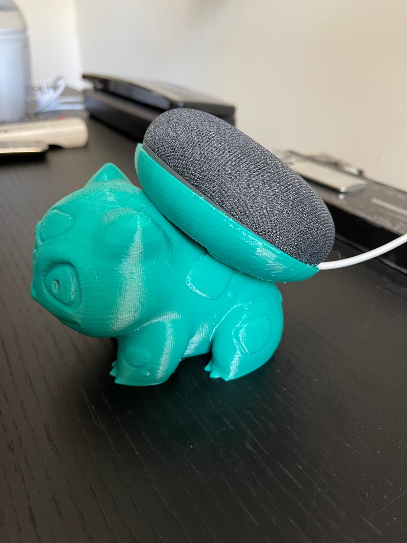 May include: A teal green 3D printed figurine of a Pokemon character, Bulbasaur, holding a gray Google Home Mini smart speaker.