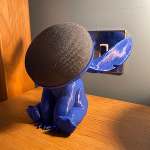 May include: A blue 3D printed figurine of Stitch from Lilo & Stitch, with a black speaker on its head. The figurine is sitting on a wooden surface.
