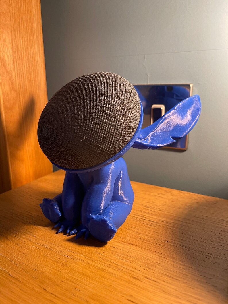 May include: A blue 3D printed figurine of Stitch from Lilo & Stitch, with a black speaker on its head. The figurine is sitting on a wooden surface.