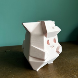 May include: A white 3D printed figurine of a cartoon character with a geometric design. The character has a smiling face with pink eyes and a pink nose.