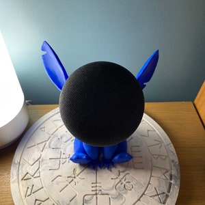 May include: A blue Stitch character figurine holding a black smart speaker. The figurine is sitting on a white circular base with a black and white marbled design.