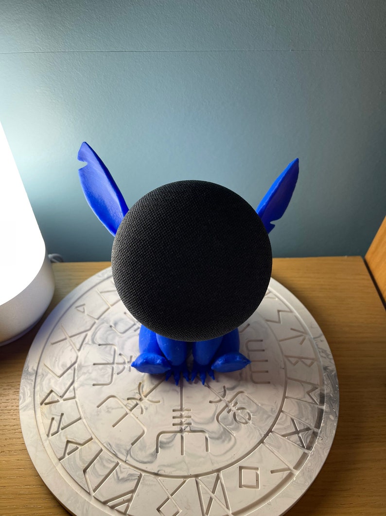May include: A blue Stitch character figurine holding a black smart speaker. The figurine is sitting on a white circular base with a black and white marbled design.