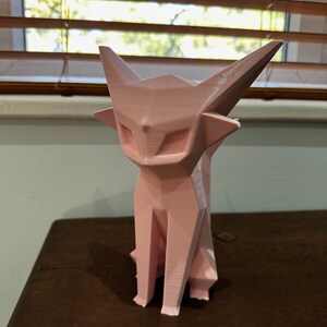 May include: A pink, geometrically-shaped cat figurine. The cat is sitting with its legs tucked underneath it. The figurine is made of a smooth, plastic-like material.