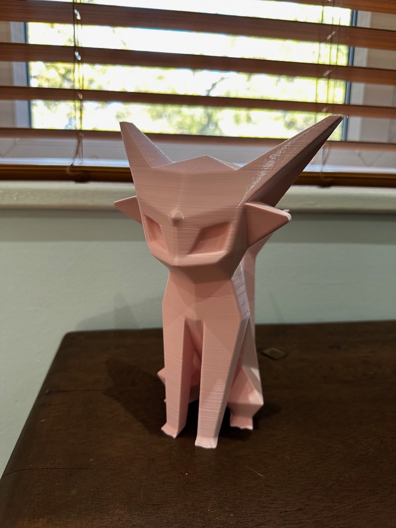 May include: A pink, geometrically-shaped cat figurine. The cat is sitting with its legs tucked underneath it. The figurine is made of a smooth, plastic-like material.
