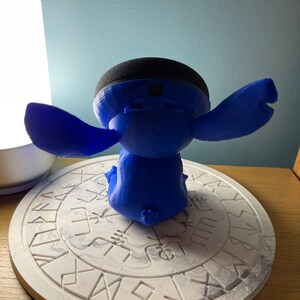 May include: A blue 3D printed Stitch character holding a black smart speaker. The character is sitting on a white circular coaster with a grey marbled design and a pattern of symbols.