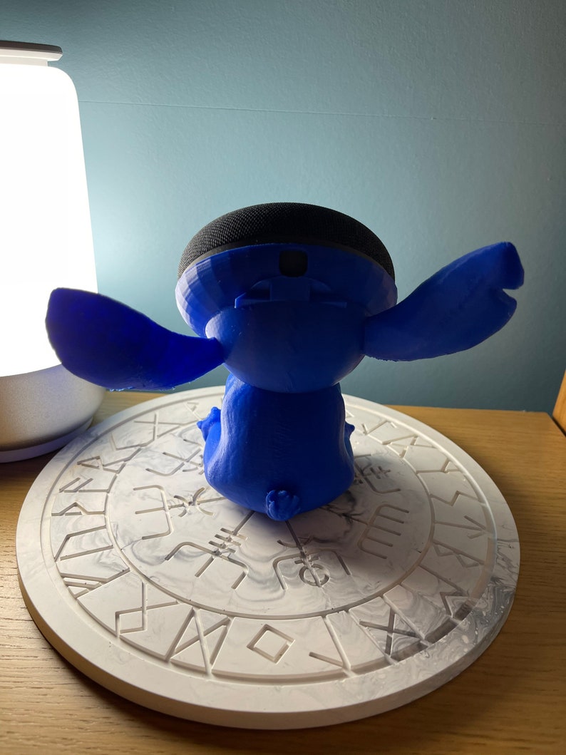 May include: A blue 3D printed Stitch character holding a black smart speaker. The character is sitting on a white circular coaster with a grey marbled design and a pattern of symbols.
