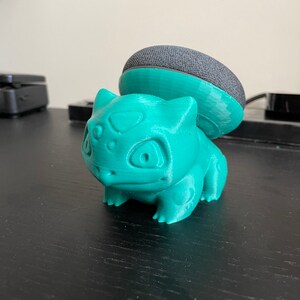 May include: A teal green 3D printed figurine of a Pokemon character, Bulbasaur, with a black and gray smart speaker on top of its head.