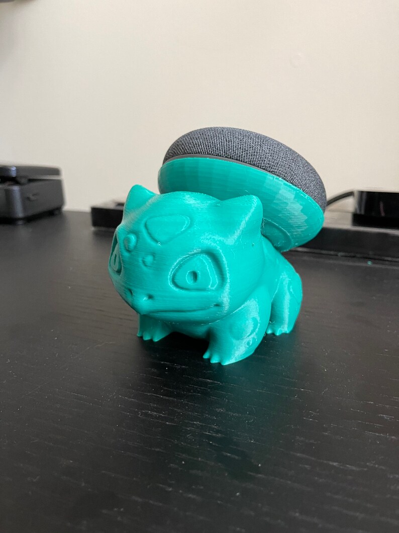 May include: A teal green 3D printed figurine of a Pokemon character, Bulbasaur, with a black and gray smart speaker on top of its head.