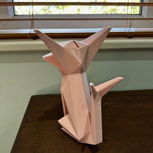 May include: A pink origami-style cat figurine made of plastic. The cat is sitting up with its ears perked up and its tail curled around its body.