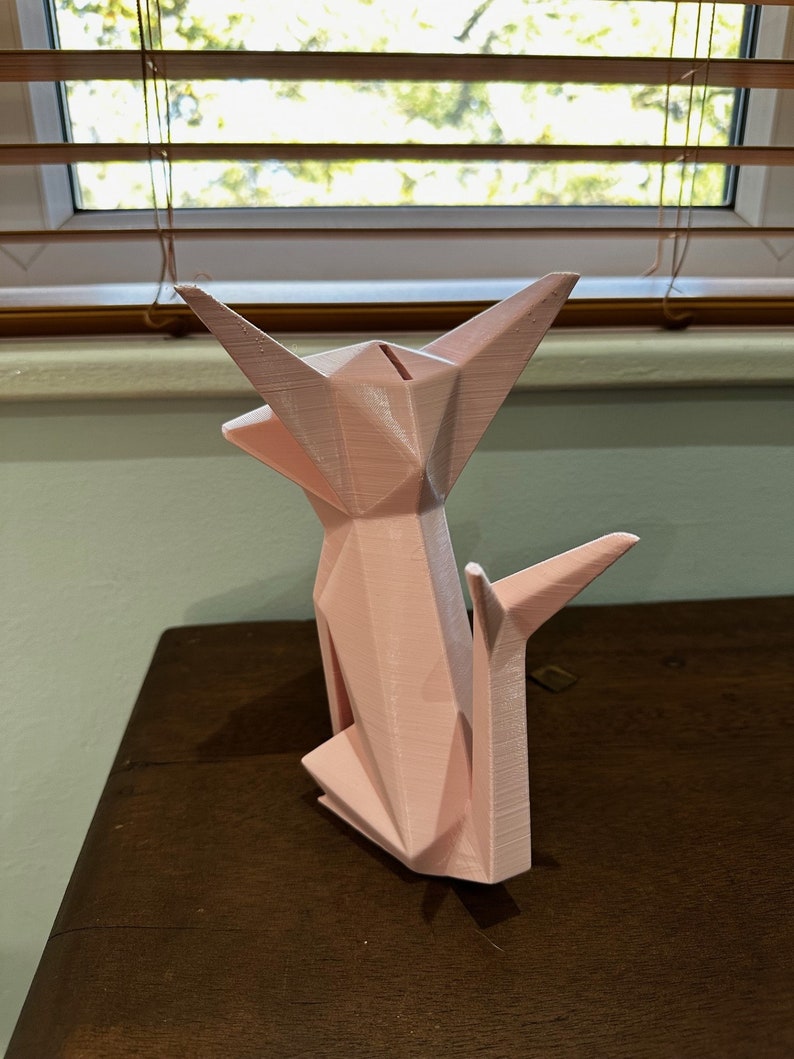 May include: A pink origami-style cat figurine made of plastic. The cat is sitting up with its ears perked up and its tail curled around its body.