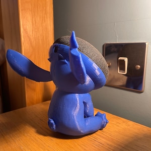 May include: A blue 3D printed figurine of Stitch from Lilo & Stitch, sitting with one arm raised. The figurine is designed to hold a smart speaker.