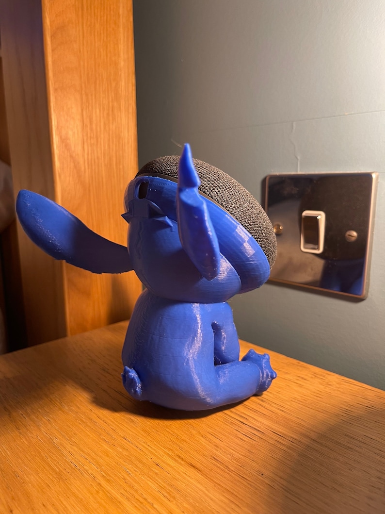 May include: A blue 3D printed figurine of Stitch from Lilo & Stitch, sitting with one arm raised. The figurine is designed to hold a smart speaker.