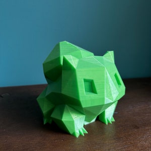 Peut inclure: Une figurine en 3D imprim&eacute;e en vert, de style low-poly, repr&eacute;sentant un personnage de Pokemon. La figurine est pos&eacute;e sur une surface en bois.