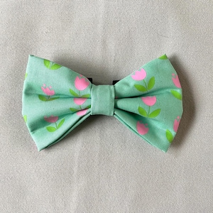 Op de afbeelding: Een mintgroene vlinderdas met een bloemmotief van roze tulpen en groene bladeren. De stoffen vlinderdas heeft een zwarte verstelbare band. Een modieus accessoire.