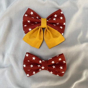 Può includere: Due papillon in tessuto con un motivo a stelle bianche su sfondo bordeaux. Un papillon è sovrapposto a un tessuto giallo senape tinta unita.