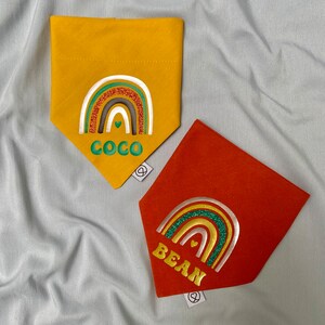 Op de afbeelding: Twee hondenbandanas met regenboogontwerpen en glitteraccenten. De gele bandana zegt "Coco" en de oranje bandana zegt "Bean".