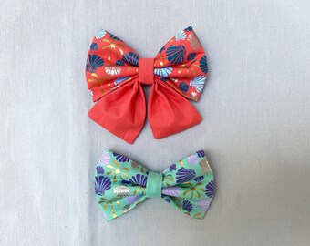 May include: Two fabric hair bows with a seashell pattern. The larger bow is red with gold accents and the smaller bow is green with gold accents.