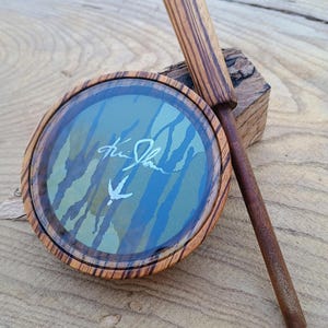May include: A wooden duck call with a camouflage design and a signature on the lid. The call is made of wood with a brown and black striped pattern.