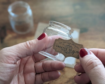 Puede incluir: Un pequeño tarro de cristal transparente con base cuadrada y cuello corto. Se está aplicando una etiqueta marrón con el texto "THE DEW DROP DISTILLERY POTION OF GREATER HEALIN premium quality" al tarro.